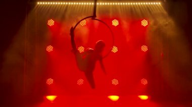 Hava akrobat kız bir çemberin üzerinde havada dönüyor ve akrobatik elementleri sergiliyor. Arka planda kırmızı ışıklar olan karanlık bir stüdyoda çekilmiş. Siluet. Yavaş çekim.