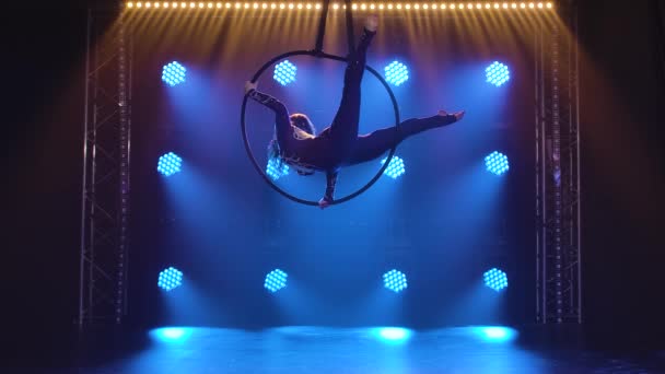 Aerial acrobat in the ring. Beautiful girl performs the acrobatic elements in the air ring. Shot in a dark studio with blue neon lights in the background. Slow motion. — Stock Video