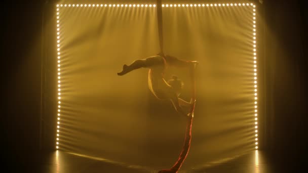 Equilibrio gimnasta estirada y girando sobre una seda roja. Silhouette niña realiza los elementos acrobáticos en el maillot apretado. Fondo ahumado oscuro y efectos de luz de neón. Movimiento lento. — Vídeos de Stock