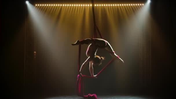Equilibrium gymnast stretched and rotating on a red silk. Silhouette young girl performs the acrobatic elements in tight leotard. Dark smoky background and neon light effects. Slow motion. — Stock Video