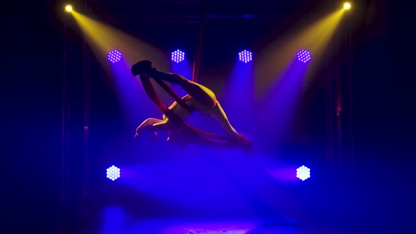 Ginasta aérea feminina realizando girar e cordel em uma seda vermelha. Espectáculo acrobático emocionante. Fundo fumegante escuro e efeitos de luz de néon azul. Movimento lento. — Vídeo de Stock