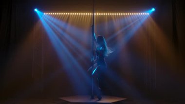 Genç bir kadının atletik fit vücudunun pilon üzerinde şehvetli danslar yapan seksi bir kadın. Mavi ışıklı karanlık bir stüdyo. Yavaş çekim.