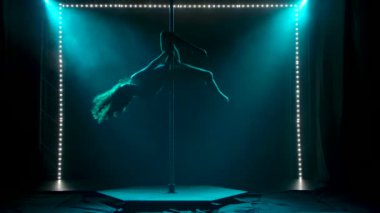 İnce kadın bir direğin üzerinde akrobatik egzersizler yapıyor. Yumuşak turkuaz ışıkta siluetler.