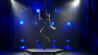 Sevimli genç bir kadın yavaş çekimde direğin üzerinde dönüyor. Stüdyoda mavi arka plan. Direk dansı..