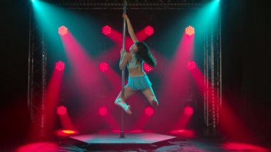 Atletik esnek dansçı, parlak kırmızı neon ışıkları olan bir stüdyoda direğin üzerinde döner. Dans eden koreografi elementleri. Vücut egzersizlerinin mükemmel performansı. Yavaş çekim.