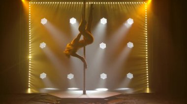 Direkte dans eden genç bir kadın, dansçı, fitness ve spor. Direk Dansı. Stüdyonun sarı ışıklarıyla siyah arka planda ince, atletik bir vücudun silueti. Yavaş çekim.