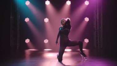 Stüdyoda plastik döşeme eğitimi alan bir kadın. Siyah arka planda mor spot ışıklarıyla hareket eden şehvetli sarışın. Yavaş çekim.