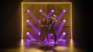 Siyah beyaz takım elbiseli üç erkek jimnastikçi bir baston çeviriyor. Erkekler mor spot ışıklarının arkasında poz veriyorlar. Yavaş çekim.