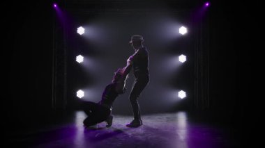 Bir grup erkek jimnastikçi dengeyi sağlamak için akrobatik hareketler sergiliyor. Siyah arka planda spot ışıkları olan iki adamın siluetleri. Yavaş çekim.