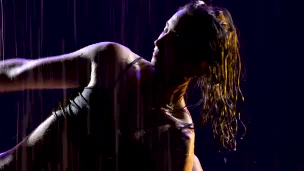 Λεπτή γυναίκα υγρή από τη βροχή ασκεί γιόγκα asanas σε ένα σκοτεινό στούντιο. Το σέξι σώμα καλύπτεται με σταγονίδια νερού που λάμπουν στο φως του στούντιο. Μαύρο φόντο. Κλείσε.. — Αρχείο Βίντεο