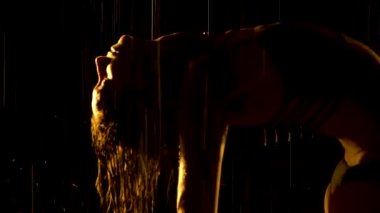 Yağmurdan ıslanmış siluet gibi ince bir kadın karanlık bir stüdyoda yoga yapıyor. Seksi vücut stüdyo sarı ışıkta parlayan su damlacıklarıyla kaplıdır. Siyah arka plan. Kapat..