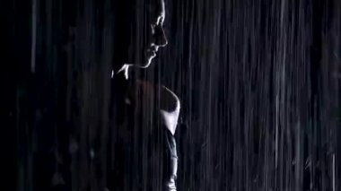 Sağanak yağmurda, genç bir kadın yoga asanası yapıyor. Stüdyo ışığı ve su damlalarında ince bir vücudun silueti. Siyah arka plan. Kapat..