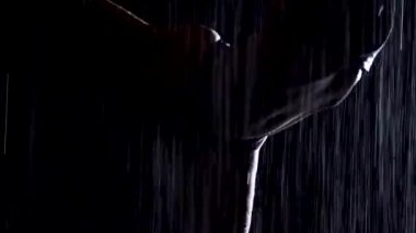 Sağanak yağmurda, genç bir kadın yoga asanası yapıyor. Stüdyo ışığı ve su damlalarında ince bir vücudun silueti. Siyah arka plan. Kapat..