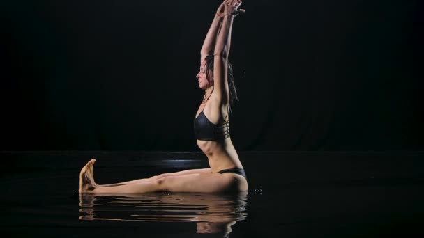 Mulher sentada na superfície da água em estúdio escuro realizando asanas para a frente dobrar de uma posição sentada. Corpo esbelto molhado brilha em luz de estúdio contra um fundo preto. Movimento lento. — Vídeo de Stock