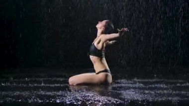 Yağmur altında yerde oturan kadın ellerini nilüfer çiçeği pozu ve huzur ve sükunet içinde katlıyor. Suyun yüzeyindeki karanlık bir stüdyoda çekilmiş. Siyah arka plan. Yavaş çekim.