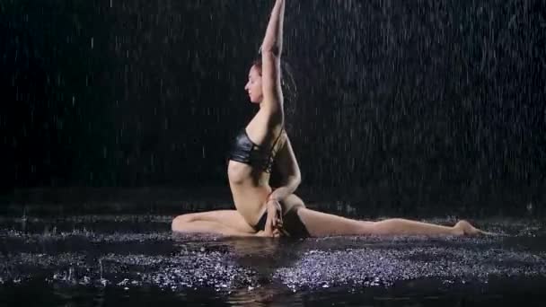 Mujer deportiva haciendo ejercicios de estiramiento y se sienta en el cordel bajo los arroyos de lluvia. Gotas de agua caen sobre el cuerpo sobre fondo negro. Movimiento lento. — Vídeos de Stock