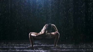 Yağmurdan sırılsıklam olmuş zayıf bir kadın karanlık bir stüdyoda egzersiz yapıyor. Seksi vücut stüdyo ışıklarında parlayan su damlacıklarıyla kaplıdır. Siyah arka plan. Yavaş çekim.