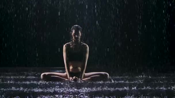 Sportvrouw doet stretching oefeningen onder de stromen van regen. Druppels water vallen op het lichaam tegen een zwarte achtergrond. Studio licht creëert glans op het oppervlak van het water. Langzame beweging. — Stockvideo