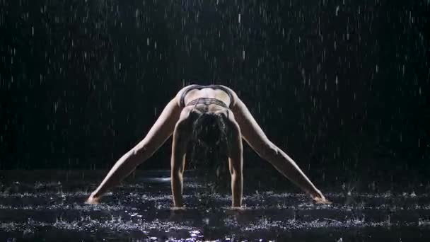 Smal kvinna våt från regnet utövar yoga asanas i en mörk studio. Den sexiga kroppen är täckt med vattendroppar som glittrar i studioljuset. Svart bakgrund. Långsamma rörelser. — Stockvideo