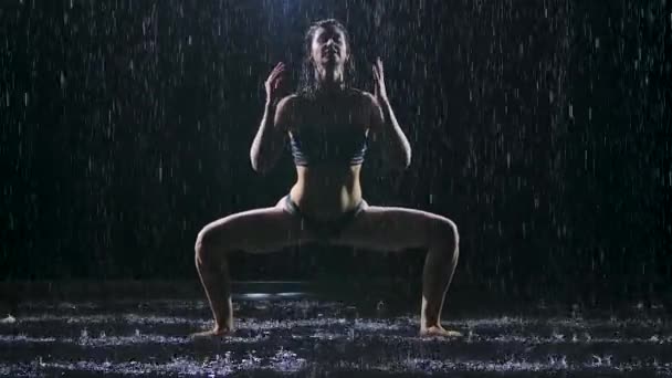 Femme mince mouillée par la pluie faisant des exercices d'étirement dans un studio sombre. Le corps sexy est recouvert de gouttelettes d'eau qui brillent dans la lumière du studio. Fond noir. Mouvement lent. — Video