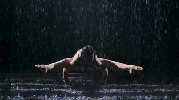 Froschpose einer jungen Sportlerin in einem dunklen Studio im Regen. Studiolicht erzeugt Schimmer auf der Oberfläche des Wassers und des Körpers. Gesunder Lebensstil. Zeitlupe. — Stockvideo