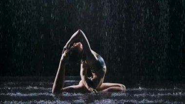 Etrafı yağmur damlalarıyla çevrili ince bir kadın tarafından yapılan yoga egzersizleri. Güvercin pozu. Seksi vücut stüdyo ışıklarında parlayan su damlacıklarıyla kaplıdır. Siyah arka plan. Yavaş çekim.