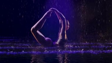 Yağmurda karanlık bir stüdyoda genç bir sporcu tarafından poz verildi. Stüdyo ışığı suyun ve vücudun yüzeyinde parıltı yaratır. Sağlıklı yaşam tarzı kavramı. Yavaş çekim.