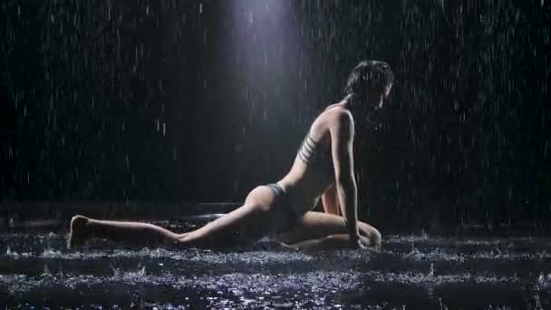由一个身材苗条的女人进行的伸展瑜伽练习，周围都是雨滴。性感的身体上布满了水滴，在演播室的灯光下闪闪发光。黑色背景。慢动作. — 图库视频影像