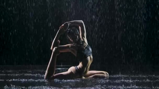 Exercices de yoga extensible effectués par une femme mince entourée de gouttes de pluie. Le corps sexy est recouvert de gouttelettes d'eau qui brillent dans la lumière du studio. Fond noir. Mouvement lent. — Video