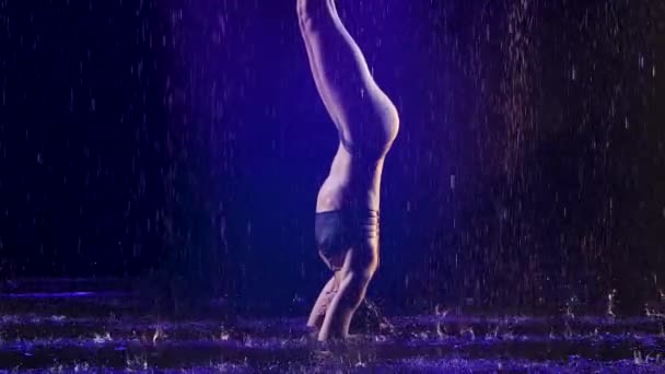 Sportieve vrouw staat in geavanceerde headstand met studio blauw licht en water regen. Vrouwelijk lichaam in druppelwater. Foto genomen in de studio op een zwarte achtergrond. Langzame beweging. — Stockvideo