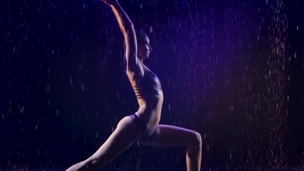 Jonge vrouw doet yoga oefeningen met studio blauw licht en water regen. Vrouwelijk lichaam in druppelwater. Foto genomen in de studio op een zwarte achtergrond. Langzame beweging. — Stockvideo