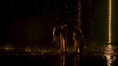 Briç pozisyonunda yoga pratiği yap. Sarı ışık, genç ve çekici bir kadının ıslak vücuduna hafifçe düşer. Vücut esnekliğinin kanıtı. Siluetli. Bardaktan boşanırcasına yağmur yağan bir stüdyo. Yavaş çekim.