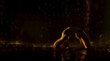 Briç pozisyonunda yoga pratiği yap. Sarı ışık, genç ve çekici bir kadının ıslak vücuduna hafifçe düşer. Vücut esnekliğinin kanıtı. Siluetli. Bardaktan boşanırcasına yağmur yağan bir stüdyo. Yavaş çekim.