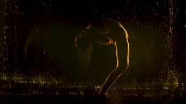 Genç bir kadın, yağmurda karanlık bir stüdyoda yoga yapıyor. Siyah arkaplan ve sarı neon ışık. Cesedin su damlalarındaki silueti. Yavaş çekim.