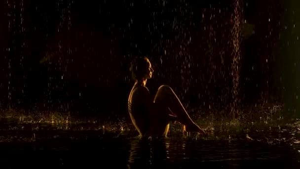 Ubhaya Padangusthasana, gespielt von einer flexiblen Frau. Der Regen prasselt auf den halbnackten Körper des Athleten. Gelbes weiches Licht in einem dunklen Studio mit schwarzem Hintergrund. Zeitlupe. — Stockvideo