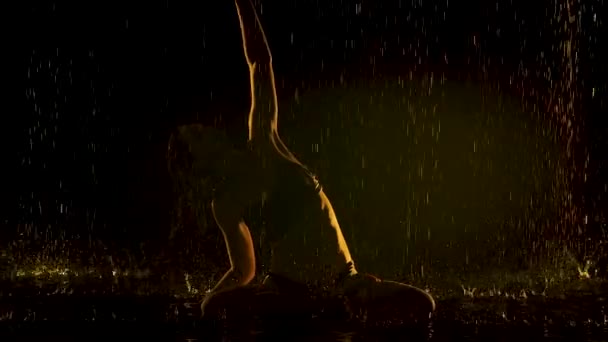 요가를 연습하는 날씬 한 여자의 실루엣이다. 빗방울이 천장에서 떨어지고 있습니다. 노란색 스튜디오 의 빛은 물의 표면과 몸에 부드럽다. 느린 동작. — 비디오
