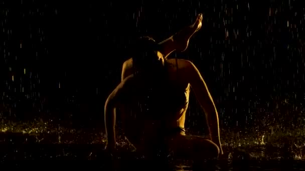 頭部の後ろの足を練習しなさい。若い女性は雨の中で暗いスタジオでヨガのアーサナを練習します。黒の背景と黄色のネオンライト。水の滴で体のシルエット。スローモーション. — ストック動画