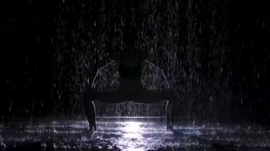 Karanlık bir stüdyoda sağanak yağmurda yoga egzersizleri yapmak. Beyaz bir spot ışığı suyun yüzeyini ve genç bir kadının vücudunu aydınlatır. Siluet. Yavaş çekim.