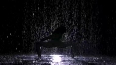 Karanlık bir stüdyoda sağanak yağmurda yoga egzersizleri yapmak. Beyaz bir spot ışığı suyun yüzeyini ve genç bir kadının vücudunu aydınlatır. Siluet. Yavaş çekim.