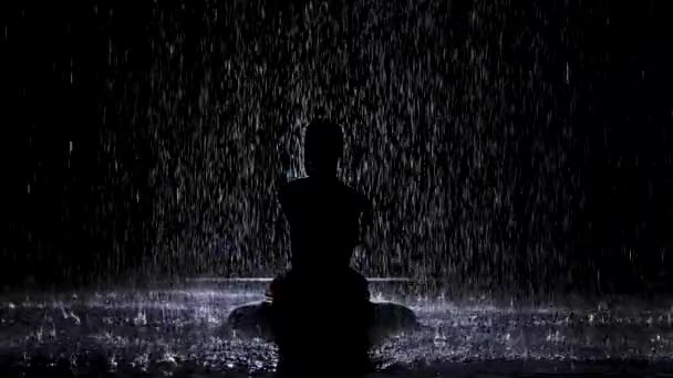 Una silueta se cierne bajo los arroyos de lluvia con las manos dobladas en una posición de loto. Fondo negro y luz de neón. Movimiento lento. — Vídeos de Stock