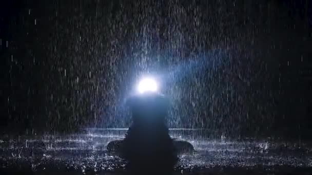 Una silueta se cierne bajo los arroyos de lluvia con las manos dobladas en una posición de loto. Fondo negro y luz de neón. Movimiento lento. — Vídeos de Stock