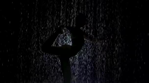 ダンサーは土砂降りの雨の中でポーズ。ネオンさわやかな白と暗いスタジオでヨガ演習を練習若い女性のぬれたボディシルエット。スローモーション. — ストック動画