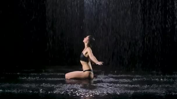 若い女性がスタジオで水の表面に座って瞑想します。女性は雨が降っているのを楽しむ。暗いスタジオで撮影。スローモーション. — ストック動画