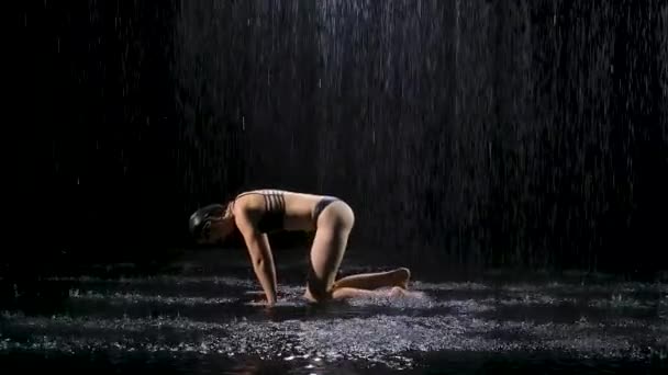 Mujer bajo la lluvia practica yoga realizando asanas. Posar hacia abajo con la pierna doblada. Cuerpo femenino apretado en gotas de lluvia. Grabado en el estudio sobre un fondo negro. Movimiento lento. — Vídeo de stock