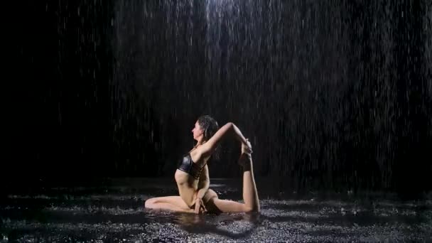 Sportovní žena pózovala pod proudem deště. Kapky vody padají na tělo na černém pozadí. Studiové světlo vytváří třpyt na hladině vody. Zpomalený pohyb. — Stock video