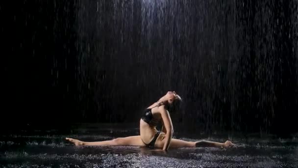 Bajo los torrentes de lluvia, una joven se sienta sobre un cordel y disfruta de un ejercicio. La luz del estudio crea brillo en la superficie del agua. Movimiento lento. — Vídeos de Stock