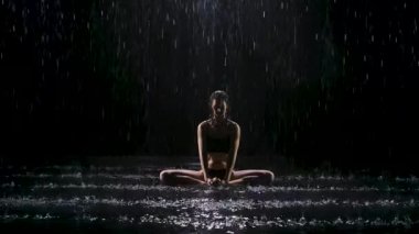 Yağmurun dereleri altında esneme egzersizleri yapan genç bir kadın. Siyah zemine su damlaları düşer. Stüdyo ışığı suyun yüzeyinde parıltı yaratır. Yavaş çekim.