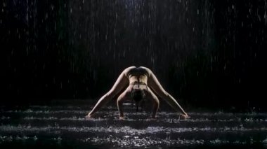 Yağmurun dereleri altında esneme egzersizleri yapan genç bir kadın. Siyah zemine su damlaları düşer. Stüdyo ışığı suyun yüzeyinde parıltı yaratır. Yavaş çekim.
