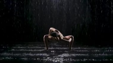 Karanlık bir stüdyoda yağmur altında yoga egzersizleri yapan bir kadın. Spot ışıkları suyun yüzeyini ve genç bir kadının vücudunu aydınlatır. Yavaş çekim.