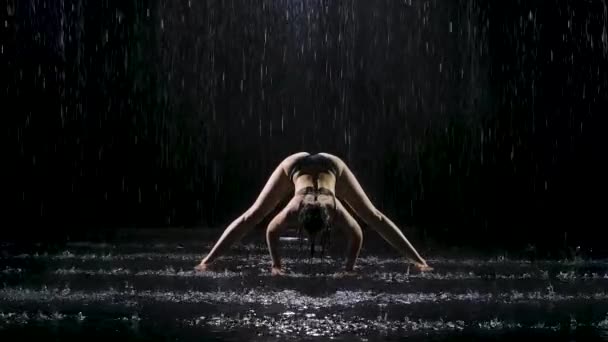 한 젊은 여자가 비가 내리는 강물 아래서 스트레칭 운동을 하고 있습니다. 몸에 떨어지는 물방울이 검은 색 바탕에 떨어진다. 스튜디오 전등은 물 표면에 작은 빛을 만들어 냅니다. 느린 동작. — 비디오
